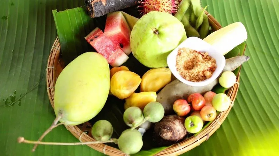A round woven bamboo basket filled with colourful tropical fruits and local produce, including green mango, sliced watermelon, guava, jackfruit pods, rambutan, edamame, and small berries. (Source: Vam Xang Orchard)